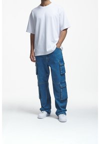 Muž v jednoduchém bílém oversized tričku, modrých cargo džínech s několika kapsami a bílých teniskách, stojící s jednou rukou v kapse.