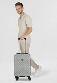 Hombre con atuendo beige tirando una maleta gris con patrón, ruedas negras y asa telescópica sobre fondo blanco.