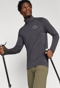 Szürke, hosszú ujjú, garbós póló logóval, raglán ujjak és sima textúra. Olívazöld nadrággal és túrázó botokkal párosítva.