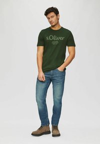 Man wearing green s.Oliver t-shirt, blue jeans, and brown lace-up boots standing with one hand in pocket on white background.