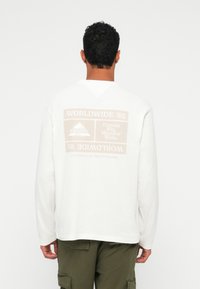 Λευκή μπλούζα με μεγάλη γραφική απεικόνιση στο πίσω μέρος που αναγράφει "WORLDWIDE '85" και "Dream Big, Wander Wide" σε μπεζ απόχρωση. Μακριά μανίκια.