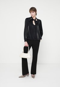 Blusa preta de cetim com um laço no pescoço, mangas longas plissadas; combinada com calças pretas e saltos com bico fino em bege; a segurar uma bolsa branca.