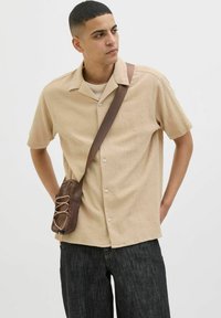 Camisa de manga corta beige con textura y diseño de botones, combinada con pantalones vaqueros oscuros y un bolso de hombro marrón compacto.