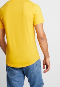 Žluté bavlněné tričko s krátkým rukávem, slim fit a kulatým výstřihem. Má hladkou texturu a jednoduchý design. Nosené s modrými denimovými džíny.