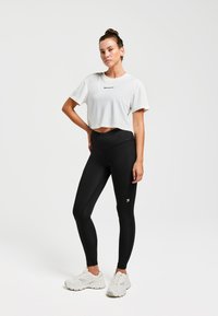 T-shirt de sport blanc coupé avec logo, associé à un legging noir taille haute avec accent logo et texture légère, complété par des baskets blanches.
