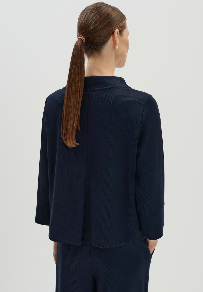 Veste bleu marine en tissu texturé, avec un col haut et une coupe légèrement décontractée. Elle possède une couture au dos et des manches arrivant au niveau des coudes.