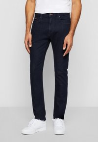 Homme portant un jean slim bleu foncé, des baskets blanches et un t-shirt blanc, debout contre un fond clair uni.