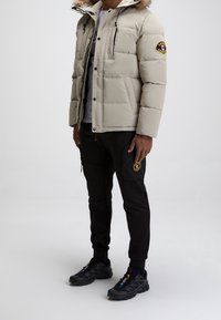 Veste rembourrée beige avec une capuche doublée en fourrure, des accents noirs et des poches zippées ; associée à un pantalon cargo noir et des chaussures de sport noires.