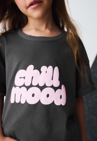 Dunkelgraues Baumwoll-T-Shirt mit dem Schriftzug "chill mood" in kräftigen, sanft rosafarbenen, abgerundeten Buchstaben. Kurze Ärmel und lockere Passform.