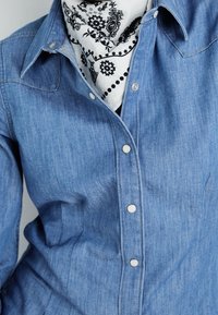 Blå denimskjorta med vita tryckknappar, spetsig krage och en svartvit mönstrad bandana synlig runt halsen.