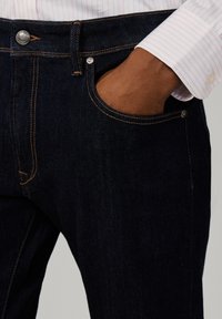 Hand, die ein weißes Langarmhemd mit pinken Streifen trägt, in die Tasche einer dunkelblauen Jeans gesteckt.