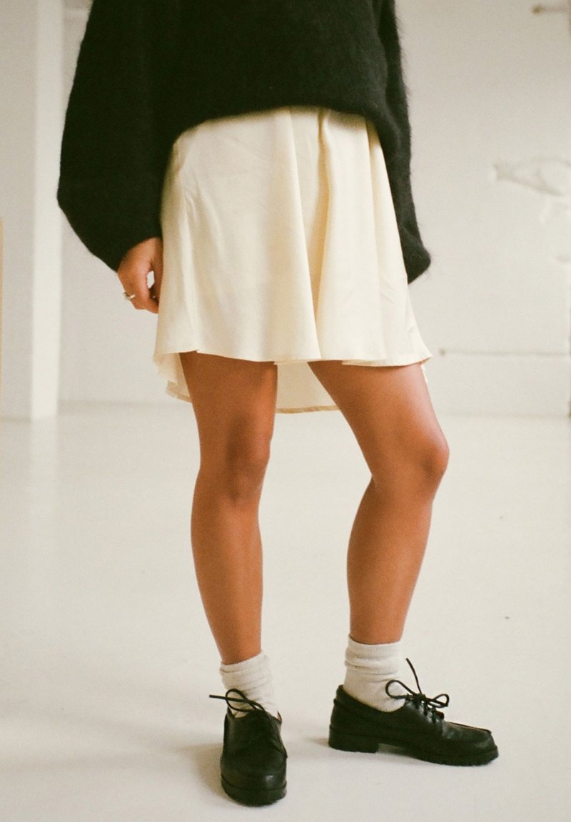 Jupe crème, longueur genoux avec des plis, associée à des chaussures noires et des chaussettes jusqu'à la cheville grises. Un grand pull noir en texture complète le look.