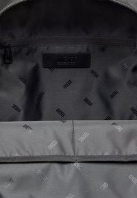 Intérieur d'un sac en tissu gris avec une texture douce, présentant une poche zippée et un patch avec le logo. Impression répétée "DIESEL" sur toute la surface.