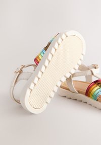 Sandales blanches avec un motif à rayures arc-en-ciel, semelle en caoutchouc texturée, bride ajustable et finition lisse.