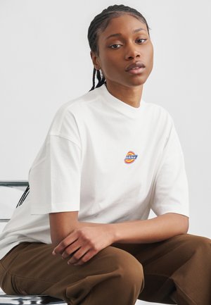 Νεαρή μαύρη γυναίκα με πλεξούδες, ντυμένη με λευκό t-shirt με το λογότυπο Dickies και καφέ παντελόνι, καθισμένη σε μεταλλική καρέκλα μπροστά σε μια απλή φόντο.