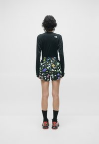 Svart langermet topp, blomster-mønstrede shorts med svart bakgrunn og sko med røde detaljer, med svarte ankelstrømper. Bakfra.