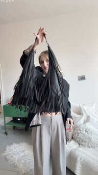 Väskan med fransar är tillverkad av svart läder och har långa, hängande fransar. Väskan hålls i en hand av en person som bär svart topp och grå byxor.