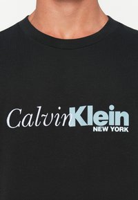 Detailní záběr na černé tričko s bílým a světle modrým logem "Calvin Klein New York" přes hruď.