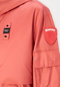 Koralen vetro jacket s kapuco, z črno oznako, rdečo vezeno oznako na rokavu in zapenjanjem na gumbke. Gladka tekstura.