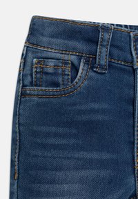 Tela de denim en azul oscuro con zonas desgastadas; presenta un bolsillo frontal, costuras amarillas y trabillas para el cinturón en la cintura.