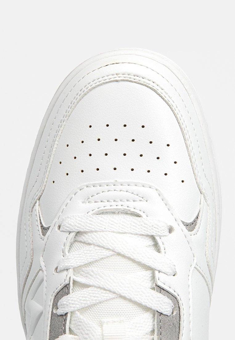 Zapatilla de cuero blanca con perforaciones en la puntera, cordones blancos y detalles de costura visibles sobre un fondo claro.