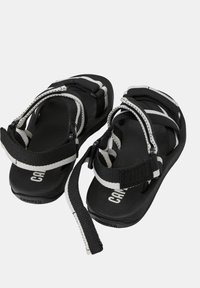 Musta-valkoiset hihnasaappaat, joissa on säädettävät Velcro-remmit ja brändätyt jalkapohjat, suunniteltu vapaamuotoiseen käyttöön.