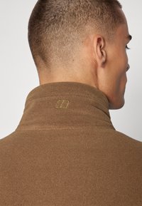 Brun høyhalset fleecegenser med en myk tekstur. Har en liten grønn brodert logo på kragen, som viser detaljerte sting.