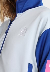 Ljusblå och mörkblå zip-upjacka med hög krage. Har en silver Reebok-logotyp och rosa detaljer på ärmarna. Mjuk tygkvalitet.