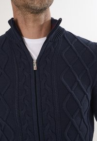 Dunkelblaues Zip-Up-Cardigan mit strukturiertem Kabelstrickmuster. Verfügt über einen hohen Kragen und einen metallischen Reißverschluss mit Markenlogo. Glatte Innenausstattung.