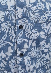 Korte mouwen overhemd in blauwe denimstructuur met witte bloem- en bladerenpatronen, met donkere knoopdetails aan de voorkant.