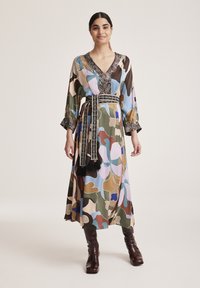 Langes, gemustertes Kleid in einer Mischung aus erdigen und pastellfarbenen Tönen, mit einem tiefen V-Ausschnitt und einem kontrastierenden Gürtel mit Quasten. Der Stoff wirkt glatt.