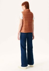 Gilet imbottito color ruggine con cuciture orizzontali, colletto alto e design senza maniche, abbinato a jeans blu scuro a campana e sneakers bianche.