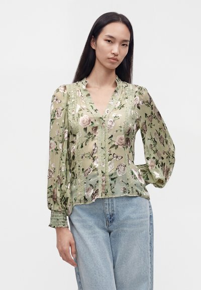 Femme portant un chemisier léger vert clair à motifs floraux transparent avec des manches longues bouffantes et un jean taille haute bleu clair sur un fond uni.