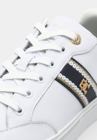 Zapatilla blanca con parte superior de cuero, franja lateral texturada en azul marino, detalles en dorado y cordones blancos. Presenta un detalle del logo en el lado.