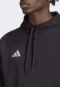 Zwarte hoodie met een zachte textuur, voorzien van een voorzak, een trekkoordkap en een wit logo met drie strepen op de linkerborst.