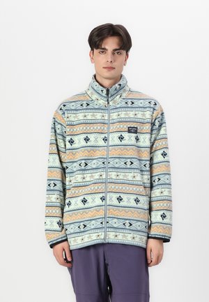 Fleece striukė su mėlynai, oranžiniais ir kremiškai baltas geometriškais raštais, aukštu apykakle ir užtrauktuku. Minkšta tekstūra.