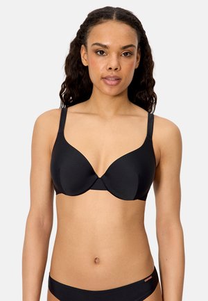 Mujer con cabello rizado y oscuro, vestida con un sujetador negro con aros y ropa interior negra a juego, de pie contra un fondo blanco liso.