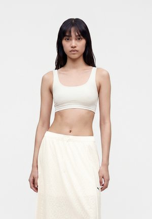Kvinde med langt sort hår iført en hvid crop top og et lysmønstret cremefarvet nederdel, stående mod en ensfarvet hvid baggrund.