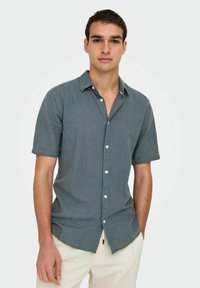 Camisa de manga corta, abotonada, en un color azul verdoso apagado, hecha de una tela texturizada. Presenta un cuello afilado y botones blancos. Diseño ligero.