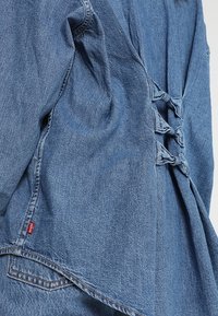 Ljusblå denimtop med lös passform, knytband i ryggen för justering och rundad nederkant. Inkluderar en liten röd etikett vid sömmen.