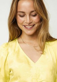 Gelbe Bluse mit floralem Muster, tiefem V-Ausschnitt und puffigenÄrmeln. Gestylt mit einer zarten Goldkette und glattem Haar.