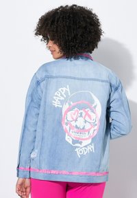 Veste en jean bleu clair présentant un graphique arrière d'un crâne avec des accents roses, le texte "HAPPY" et "TODAY", et des détails de bord frangés en rose.