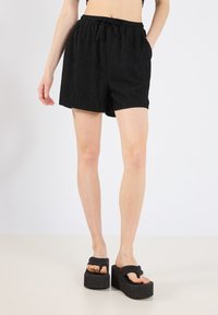Black, textured, ribbed shorts with an elastic drawstring waistband and side pockets, paired with thick-soled black sandals.