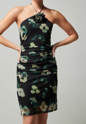 Femme portant une robe noire cintrée à col halter avec des motifs floraux verts et beiges et un détail fleur en tissu au niveau de l'encolure.