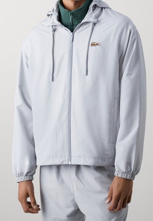 Homme portant une veste à capuche gris clair zippée avec poignets élastiques et petit logo crocodile sur la poitrine, par-dessus un haut vert à col zippé.