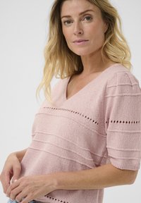 Svetlo roza pletena majica s V-izrezom in kratkimi rokavi, horizontalne črte z majhnimi izrezanimi detajli. Mehka tekstura in sproščen kroj.