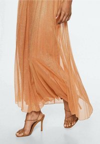 Ferskenfarvet maxi kjole i gennemsigtigt stof med en subtil glans. Har flydende lag og en sideslids, parret med nude højhælede sandaler.
