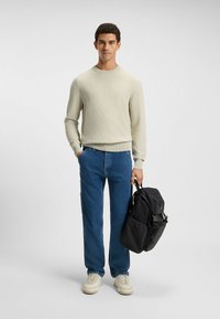 Beige strukturierter Strickpullover, blaue gerade Jeans, cremefarbene Sneaker und ein schwarzer Stoffrucksack mit Reißverschlüssen vor einem neutralen Hintergrund.
