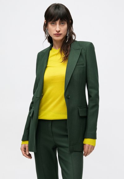 Tmavozelené sako s prispôsobeným strihom, vykrojenými klopami a jedným gombíkom na zapínanie, skombinované s žiarivým žltým svetrom.