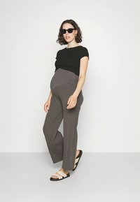 Grūtniece, tērpusies melnā topā, pelēkās ribotās grūtnieču biksēs, bēšās sandales un melnas saulesbrilles, pozē uz vienveidīga baltā fona.